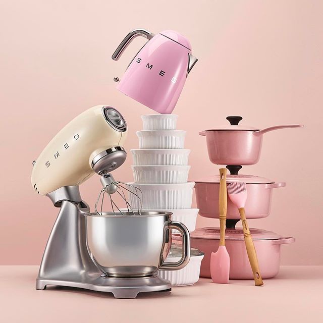 A collection of pastel-colored SMEG and Le Creuset kitchenware on a pink background.