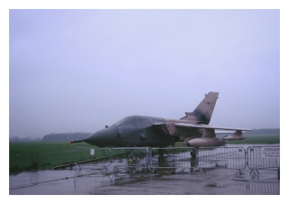 A camouflaged fighter jet sits on a rainy airfield, covered with a protective tarp, under a heavy, overcast sky.
