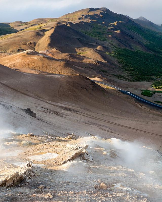 Scenic mountain landscape with a road and a car passing through a geothermal area, conveying a sense of adventure and exploration.