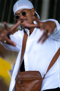 A stylish man in sunglasses and a white shirt strikes a pose with a messenger bag in an urban setting.