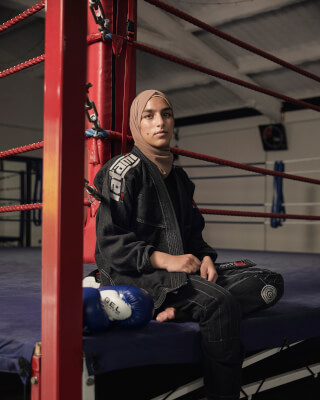 A young woman in a hijab and martial arts uniform sits in a boxing ring, exuding strength and confidence.