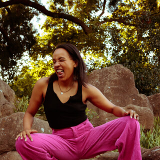 A woman in pink pants and a black top is crouching on rocks in a park with a joyful expression.