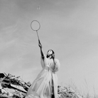 A woman in a white robe and mask hits a shuttlecock with a badminton racket outdoors in a black and white image.