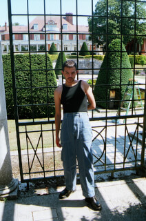 A man wearing a tank top and blue pants poses in front of a wrought iron gate in a garden.