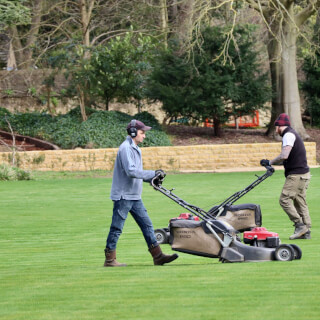 Two men mow a large green lawn with professional lawn mowers in a park setting.
