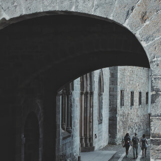 Three people walk through a stone archway in an old European town, enjoying the historic architecture and cool shade.
