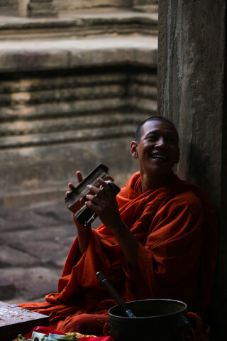 A monk smiles while holding a book, with a prayer bowl nearby in an ancient temple setting.