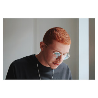 A redhead man wearing glasses and a chain necklace in a modern indoor setting.