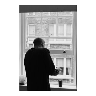 A man looks out of his apartment window at the brick buildings while smoking a cigarette. 