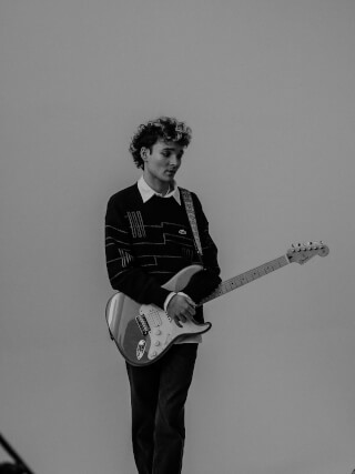 A young man with curly hair is playing an electric guitar in a black and white studio shot.