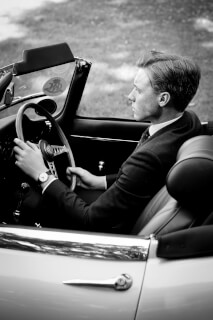 A man in a suit drives a classic car, looking ahead with a serious expression in this black and white shot.