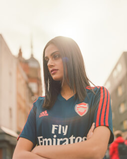 A woman in an Arsenal soccer jersey stands confidently in a city street with her arms crossed.