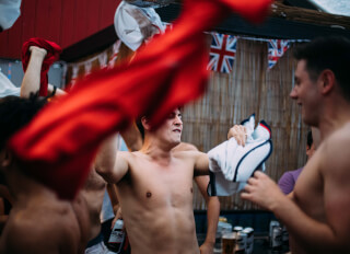 A group of shirtless men celebrate outdoors with British flags and beer at a lively gathering.
