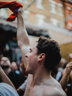 A shirtless man cheers with a crowd, raising a red and blue cloth in celebration.