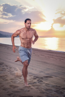 A fit man runs shirtless on a sandy beach at sunrise for fitness, health or sport campaign visuals.
