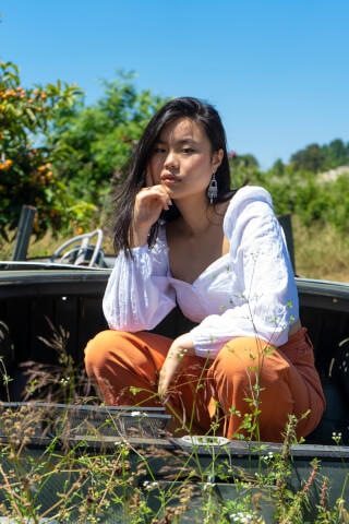 A young woman in a white top and orange pants poses outdoors in natural light.