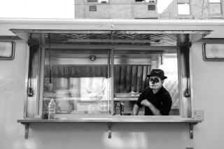A man with face paint stands in a food truck, serving customers on a city street.