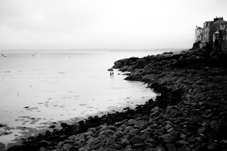 A black and white shot of two people strolling along a rocky beach with buildings on the cliffside.