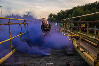 A woman poses with a purple smoke grenade on an industrial bridge for a fashion editorial.