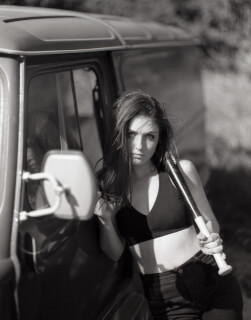 A woman with a baseball bat strikes a confident pose leaning against a car in black and white.