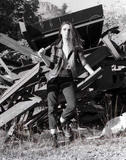 A young woman poses with a baseball bat in a black and white image in a junkyard.