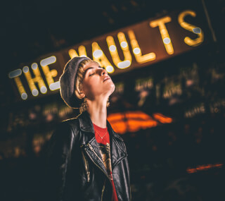 A woman in a leather jacket and beret poses at night in front of The Vaults sign.