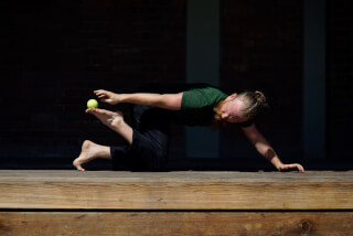 A bearded man balancing a tennis ball on his foot while practicing yoga on a wooden deck in the sunlight.