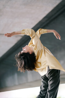 A young woman in a golden shirt gracefully dances under an urban underpass, her body arched in an elegant pose.