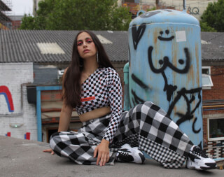 A young woman in a checkerboard outfit poses on a rooftop with an urban backdrop for a fashion-forward editorial shot.