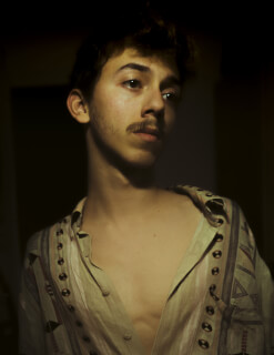 Portrait of a young man with a mustache wearing a patterned shirt, looking off to the side.