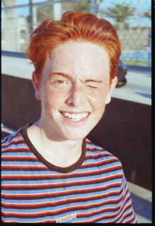 A cheerful young man with red hair and freckles winks and smiles in a candid outdoor portrait.