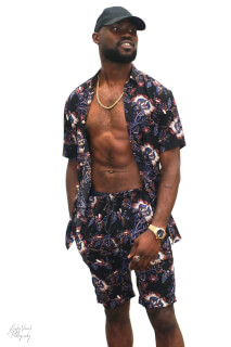 A stylish man poses in a floral print shirt and shorts, accessorized with a gold chain and watch.