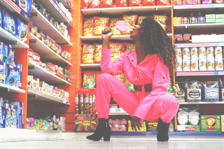 A woman in a pink jumpsuit crouches in a store aisle, enjoying a lollipop.