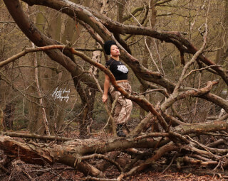 A stylish woman with a turban and camouflage pants poses in a forest setting.