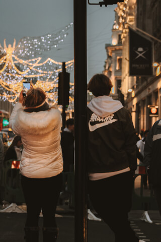 People enjoy the Christmas lights on a city street at night, capturing the festive scene with their phones.