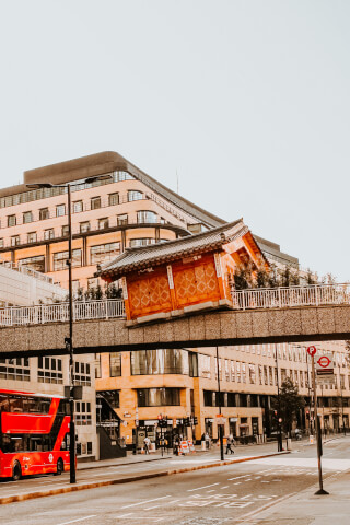 A cityscape featuring a distinctive architectural building over a walkway with a red double-decker bus passing by on a city street.