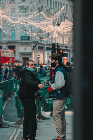 Two masked young men are standing on a city street in front of holiday lights with a crowd of people in the background.