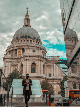 A man stands in front of St. Paul's Cathedral in London, with a red double-decker bus and black cab visible.