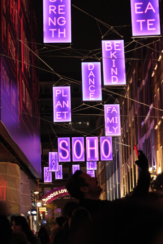 A lively street scene at night features illuminated signs spelling out positive words like 'Soho' in a vibrant urban setting.