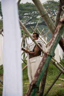 A man wearing a loincloth dries clothes on a bamboo frame in a rural setting.