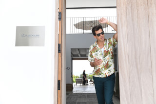 A man in a tropical shirt leans in a doorway of a modern house with a Lexus sign, enjoying a relaxed and cheerful moment.