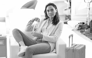 Gal Gadot sits in an airport lounge holding a Smartwater bottle in a black and white shot.