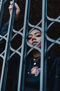 A stylish woman with jewelry and a Lakers sweatshirt poses behind a gate for a striking portrait.