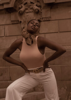 A stylish woman poses on stairs in a headscarf, top, belt and white pants.