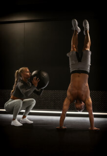 A fit woman works with a medicine ball as a man does a handstand in a gym.