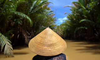 A person wearing a traditional Asian conical hat travels down a river surrounded by tropical vegetation.