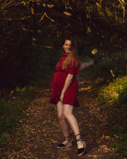 A woman in a red dress smiles while standing on a path in a lush, green forest.