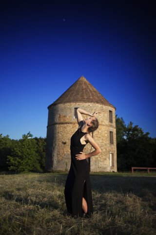 A woman in a black dress poses gracefully in a field with a stone windmill under a clear blue sky.