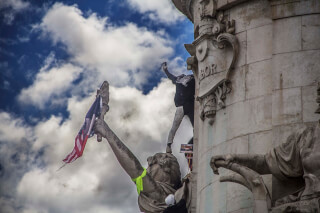 A statue is being scaled by protesters holding an American flag, showcasing intense political activism.