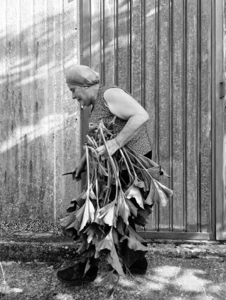 A senior woman in black and white carries a bundle of leafy greens outdoors.
